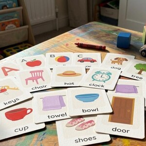 May include: A collection of flashcards with illustrations and words, including "keys," "chair," and "clock." A red crayon, wooden blocks, and a toy car are also visible. The scene suggests a learning environment for children.