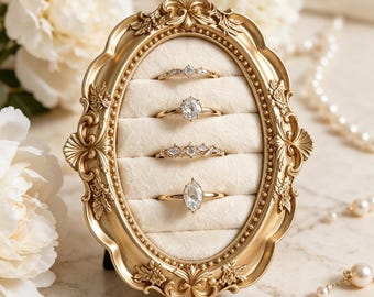 May include: An oval gold-toned frame with ornate scrollwork details containing a cream-colored velvet insert with four horizontal slots. Four gold rings with clear stones are displayed in the slots, featuring various cuts including marquise and oval.