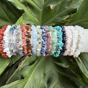 May include: A collection of gemstone chip bracelets in various colors, including red, white, brown, blue, green, and black. The bracelets are displayed on a white cylindrical surface against a backdrop of large green leaves. Each bracelet is made of small, irregular-shaped stones.