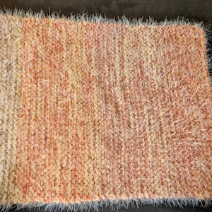 Op de afbeelding: Een rechthoekig, handgebreid tapijt met een getextureerd oppervlak. Het tapijt heeft een kleurenpalet van beige, perzik en oranje tinten. De randen zijn afgewerkt met een franje van wit garen. Dit gezellige tapijt is geschikt voor woondecoratie.