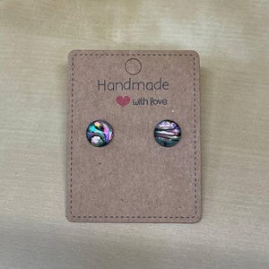 Puede incluir: Un par de pendientes de botón de concha de abulón iridiscente sobre una tarjeta marrón. La tarjeta dice "Handmade with love" en texto negro. Los pendientes tienen forma redonda y muestran una mezcla de colores, incluyendo verde, morado y azul.