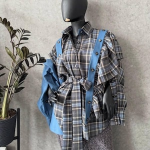 May include: A mannequin displays a plaid shirt with blue detailing and braces. The shirt has a grey, blue, and white check pattern, with a matching tie belt. The mannequin is wearing a grey skirt and black gloves.