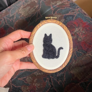 May include: An oval embroidery hoop featuring a white fabric background with a black cat silhouette stitched in the center. The hoop has a textured, wood-grain appearance and a small metal tightening screw at the top for securing the fabric.