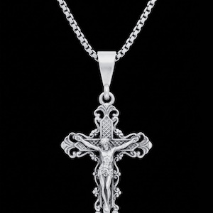 May include: Silver-toned crucifix pendant necklace. The pendant features a detailed depiction of Jesus on the cross, with ornate scrollwork. The necklace has a box chain and a simple clasp, set against a black background.