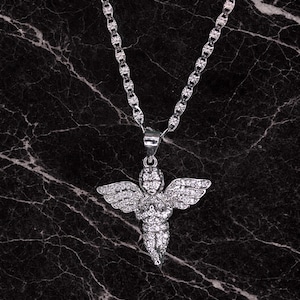 May include: A silver-colored necklace with a pendant shaped like an angel. The angel pendant has outstretched wings and is covered in small, sparkling stones. The necklace chain has a textured design, and the background is a dark marble.