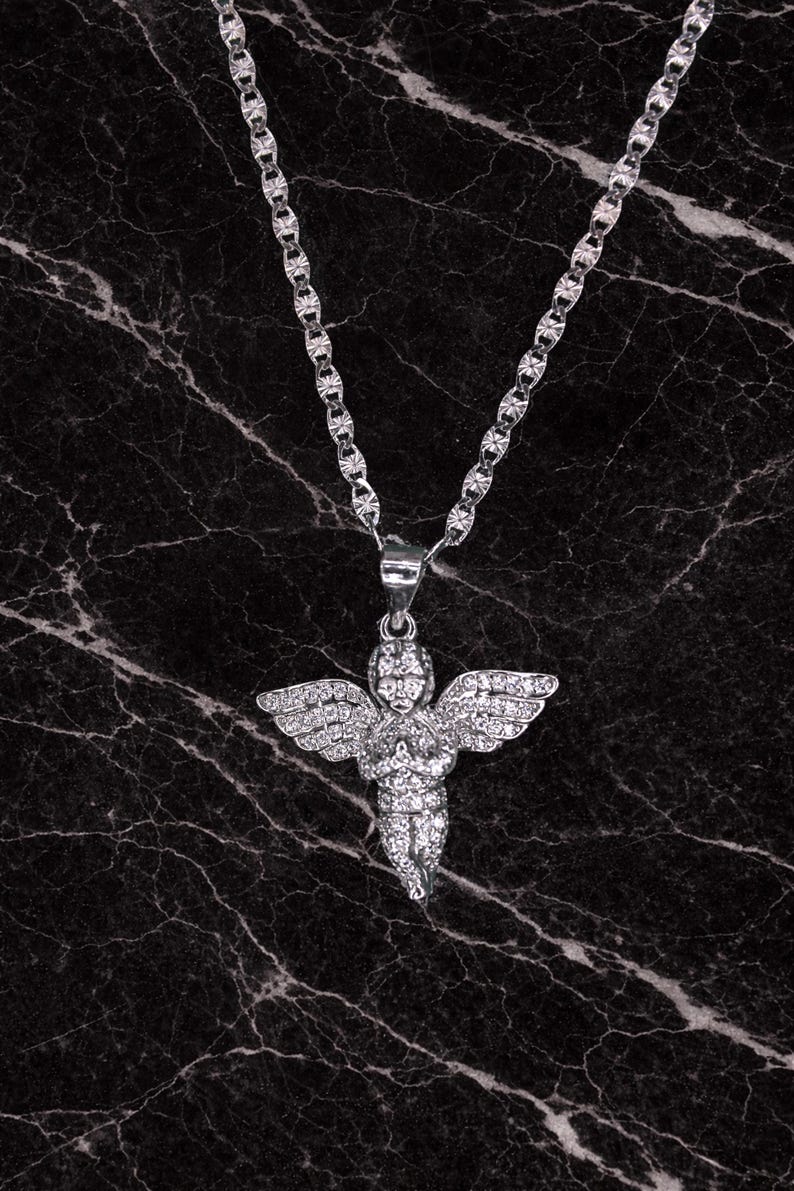 May include: A silver-colored necklace with a pendant shaped like an angel. The angel pendant has outstretched wings and is covered in small, sparkling stones. The necklace chain has a textured design, and the background is a dark marble.