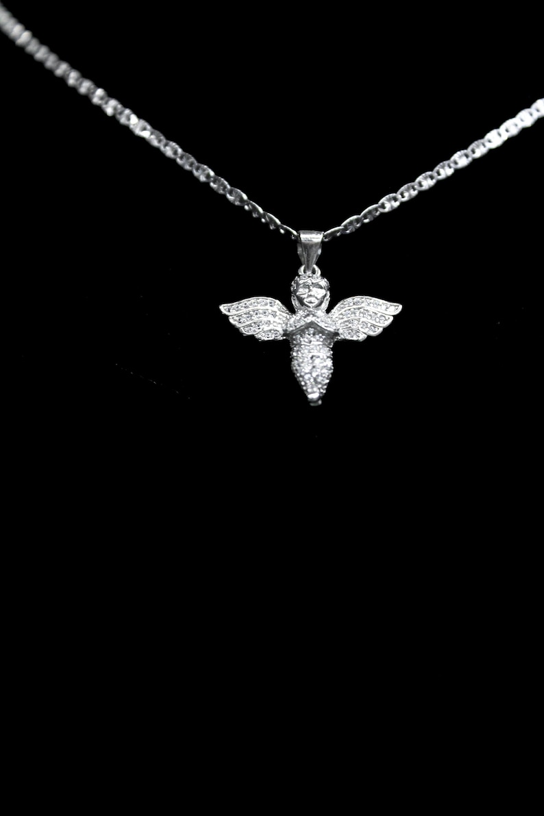 May include: A silver necklace with a pendant shaped like an angel. The angel pendant has outstretched wings and is covered in small, sparkling stones. The necklace chain is also silver and has a delicate, linked design. The background is black.