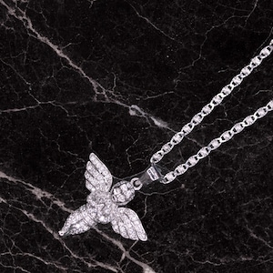 May include: A silver-colored necklace with a pendant shaped like an angel with outstretched wings. The pendant and chain are set against a dark, marbled background. The necklace is a jewelry item.