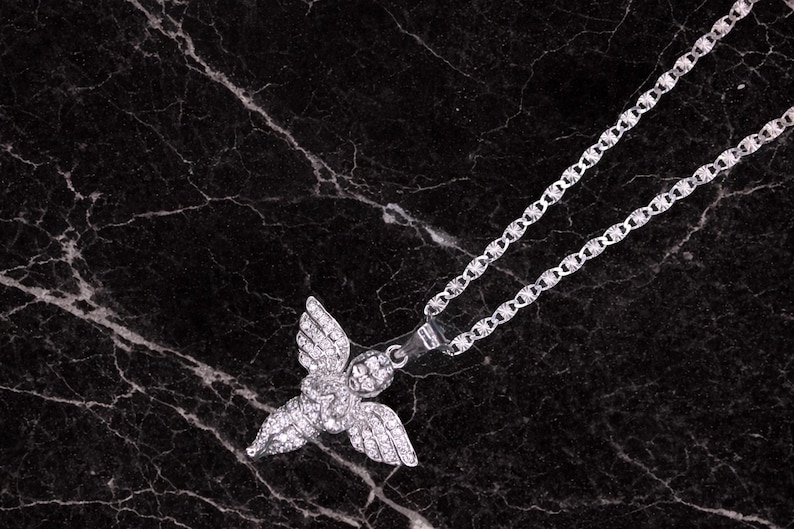 May include: A silver-colored necklace with a pendant shaped like an angel with outstretched wings. The pendant and chain are set against a dark, marbled background. The necklace is a jewelry item.