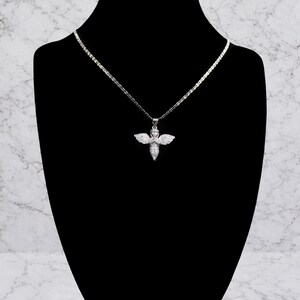 May include: A silver-toned necklace with a pendant shaped like an angel. The angel pendant has outstretched wings and is adorned with small, sparkling stones. The necklace is displayed on a black mannequin against a marble-patterned background.