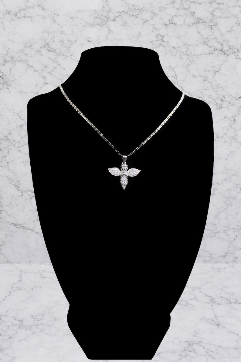 May include: A silver-toned necklace with a pendant shaped like an angel. The angel pendant has outstretched wings and is adorned with small, sparkling stones. The necklace is displayed on a black mannequin against a marble-patterned background.