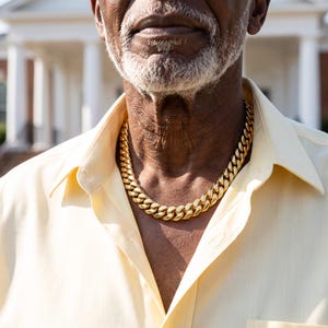 Mens Miami Cuban link chain in 18K gold plated stainless steel, worn on the neck of a man in his seventies in bright daylight.