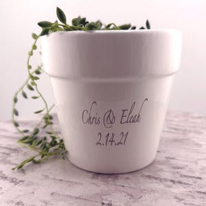 May include: A white ceramic planter with the names "Chris & Eleah" and the date "2.14.21" in black script. The planter holds a trailing green plant. The planter is on a light-colored surface.