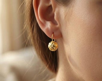 Pode incluir: Um brinco pendente de ouro com um pingente esférico que apresenta um padrão texturizado em forma de estrela. O brinco está suspenso por um gancho de arame curvo simples e exibe um acabamento metálico polido que reflete a luz.