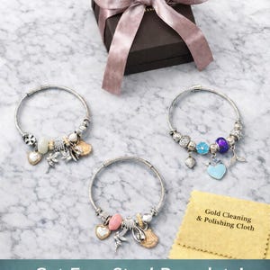 Pode incluir: Tr&ecirc;s pulseiras de berloques prateadas com v&aacute;rias contas e pingentes, incluindo conchas, cora&ccedil;&otilde;es e uma fada. Uma caixa de presente marrom escura com uma fita malva e um pano amarelo com a etiqueta Gold Cleaning & Polishing Cloth.