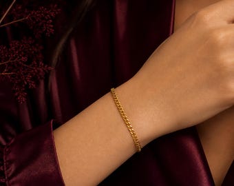 Pode incluir: Uma pulseira de corrente dourada com fecho lagosta, com 4 mm de largura. A pulseira é exibida em um pulso contra um fundo bordô escuro. A imagem destaca o design e o fecho da pulseira.