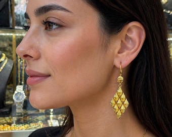 Pode incluir: Close-up de uma mulher usando brincos e colar de ouro. Os brincos têm um design em forma de diamante com detalhes intrincados. O colar é uma corrente fina. A mulher está vestindo uma blusa preta.