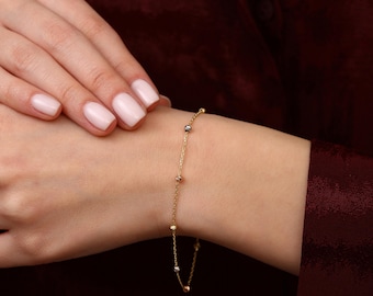 Pode incluir: Uma delicada pulseira de ouro com pequenas contas espaçadas uniformemente em ouro, prata e ouro rosa. A pulseira está em uma corrente fina e é usada no pulso. O fundo é de cor vermelho escuro sólido.