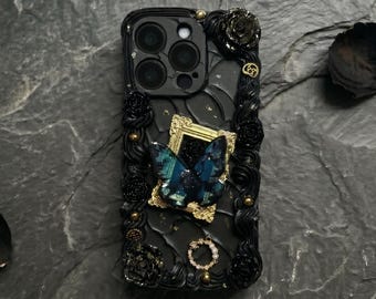 Op de afbeelding: Zwarte telefoonhoes met sierlijke details, met een goudomlijnde vlinder en rozenaccenten. De hoes heeft een textuurontwerp met gouden versieringen en een cirkelvormig juweel. De vlinder heeft blauwgroene iriserende vleugels.