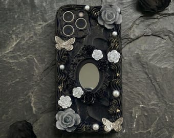Op de afbeelding: Zwarte telefoonhoes met een decoratief ontwerp met grijze en witte rozen, zilveren vlinders en imitatieparels. De hoes heeft een spiegel in het midden en is versierd met gouden accenten. De hoes is ontworpen voor een smartphone.