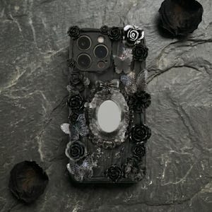 May include: Black phone case with a decorative design featuring roses, butterflies, and an oval mirror. The case is embellished with black floral accents and butterfly details, creating a gothic aesthetic.