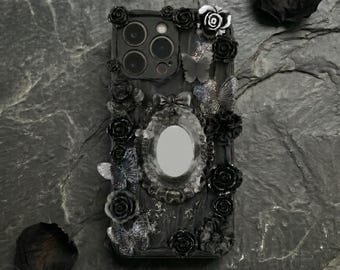 Op de afbeelding: Zwarte telefoonhoes met een decoratief ontwerp met rozen, vlinders en een ovale spiegel. De hoes is versierd met zwarte bloemenaccenten en vlinderdetails, wat een gotische esthetiek creëert.