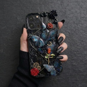 Puede incluir: Funda de teléfono negra con un diseño gótico con mariposas azules, rosas rojas y negras y pequeños engranajes. La funda tiene un fondo oscuro texturizado. La funda del teléfono la sostiene una persona con esmalte de uñas negro.
