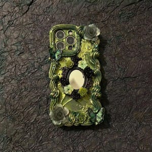 Op de afbeelding: Een limoengroene telefoonhoes met een centrale ovale spiegel omringd door licht- en donkergroene bloemversieringen, golvende verhoogde texturen en twee vlinderaccenten. De hoes heeft een uitsparing voor een drievoudig camerasysteem.