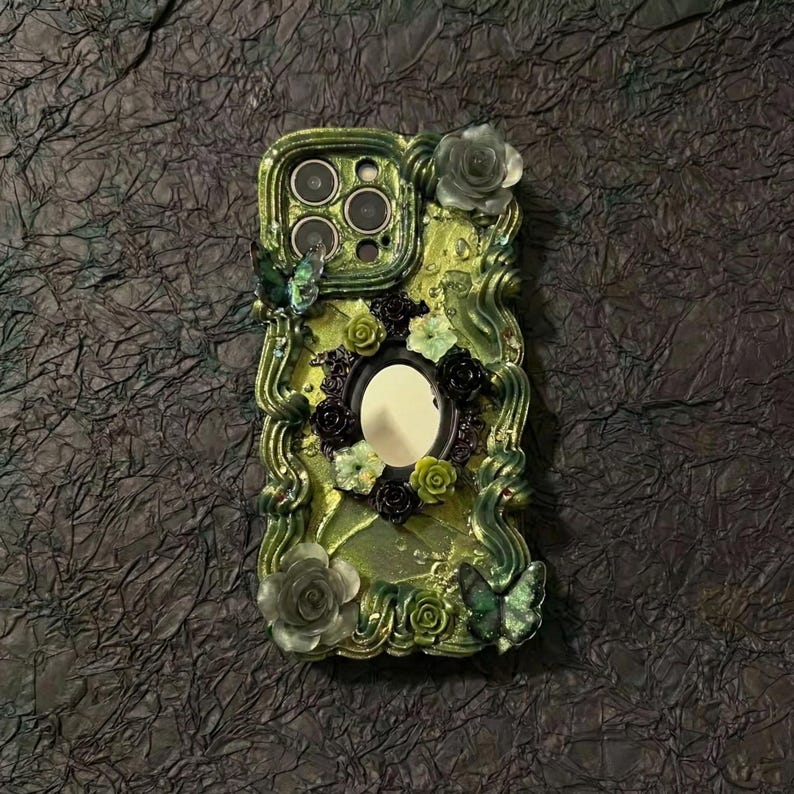 Op de afbeelding: Een limoengroene telefoonhoes met een centrale ovale spiegel omringd door licht- en donkergroene bloemversieringen, golvende verhoogde texturen en twee vlinderaccenten. De hoes heeft een uitsparing voor een drievoudig camerasysteem.