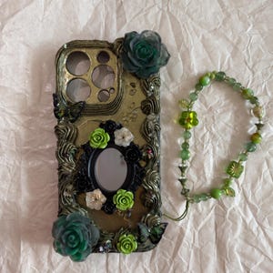Op de afbeelding: Een goudkleurig telefoonhoesje versierd met donkergroene en limoengroene harsrozen, zwarte vlinderaccenten en een centrale ovale spiegel. Een bijpassend groen kralenpolsbandje met een bloembedel is aan de zijkant bevestigd.