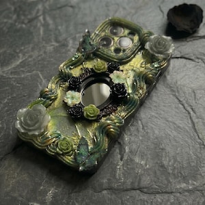 Op de afbeelding: Een decoratief telefoonhoesje met een textuur van gemarmerd groen en goud met verhoogde, wervelende accenten. Het is versierd met kleine, driedimensionale zwarte, witte en groene rozen, samen met twee iriserende vlinderfiguren en een centrale ovale spiegel.