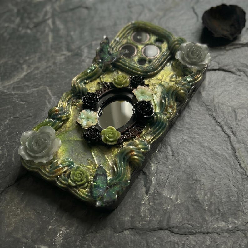 Op de afbeelding: Een decoratief telefoonhoesje met een textuur van gemarmerd groen en goud met verhoogde, wervelende accenten. Het is versierd met kleine, driedimensionale zwarte, witte en groene rozen, samen met twee iriserende vlinderfiguren en een centrale ovale spiegel.