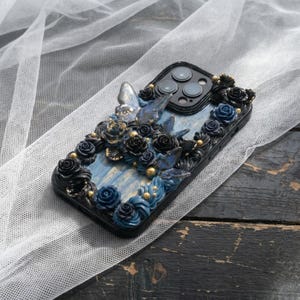 May include: Black phone case with a blue and gold floral design. The case features sculpted roses, butterflies, and gold accents. The background is a light blue with gold streaks.