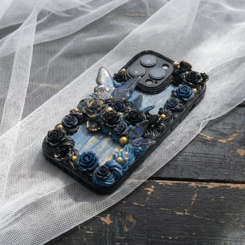 May include: Black phone case with a blue and gold floral design. The case features sculpted roses, butterflies, and gold accents. The background is a light blue with gold streaks.