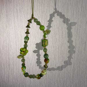 Op de afbeelding: Een kralen telefoonhanger of koord met diverse groene kralen in verschillende vormen en maten, waaronder ronde, gefacetteerde en bloemvormige glaskralen, geregen aan een dun groen koord met kleine metalen accenten.