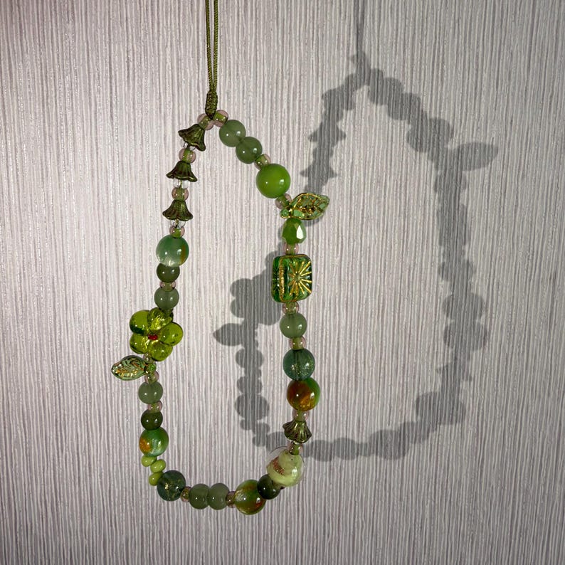 Op de afbeelding: Een kralen telefoonhanger of koord met diverse groene kralen in verschillende vormen en maten, waaronder ronde, gefacetteerde en bloemvormige glaskralen, geregen aan een dun groen koord met kleine metalen accenten.