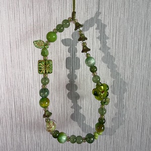 Op de afbeelding: Een kralenlus-hanger met diverse tinten groene kralen, waaronder ronde glazen bollen, een vierkante kraal met een gouden sterpatroon, een blaadjesvormige bedel, kleine klokvormige metalen tussenstukjes en een grote doorzichtige groene bloem.