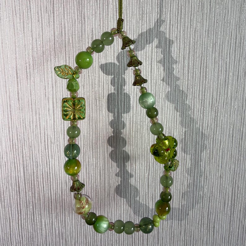 Op de afbeelding: Een kralenlus-hanger met diverse tinten groene kralen, waaronder ronde glazen bollen, een vierkante kraal met een gouden sterpatroon, een blaadjesvormige bedel, kleine klokvormige metalen tussenstukjes en een grote doorzichtige groene bloem.
