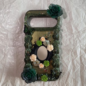 Op de afbeelding: Een rechthoekig telefoonhoesje met een donkergroene, getextureerde, rankachtige rand met twee grote donkergroene rozen in de hoeken. Het midden bevat een kleine ovale spiegel omringd door kleine cr&egrave;mekleurige en lichtgroene bloemetjes.