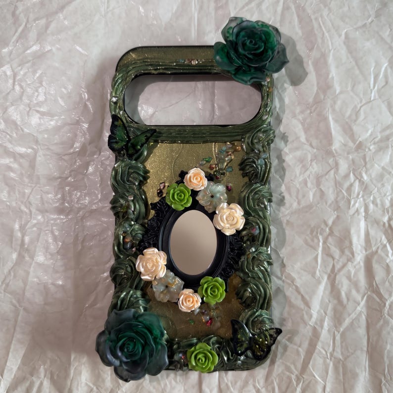 Op de afbeelding: Een rechthoekig telefoonhoesje met een donkergroene, getextureerde, rankachtige rand met twee grote donkergroene rozen in de hoeken. Het midden bevat een kleine ovale spiegel omringd door kleine cr&egrave;mekleurige en lichtgroene bloemetjes.