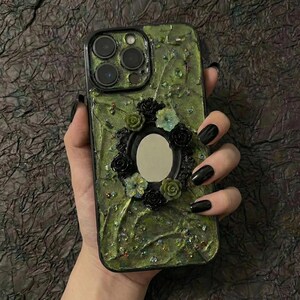 Op de afbeelding: Een smartphonehoesje met een textuurrijke olijfgroene basis, voorzien van een centrale ovale spiegellijst omringd door kleine zwarte en groene bloemversieringen en verspreide iriserende glitteraccenten op het oppervlak.