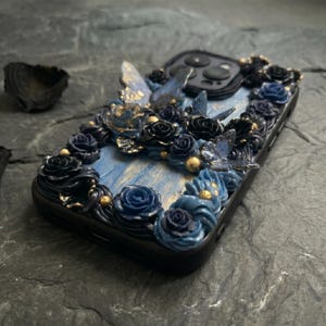 May include: Black phone case decorated with blue roses, butterflies, and gold accents. The case has a blue and gold marbled background. The design is three-dimensional and detailed, creating a unique and artistic look.