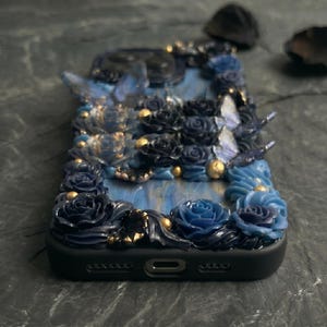 May include: A black phone case adorned with sculpted blue roses, butterflies, and gold accents. The case features a marbled blue background and is intricately detailed with a floral and butterfly design. The case has a glossy finish.