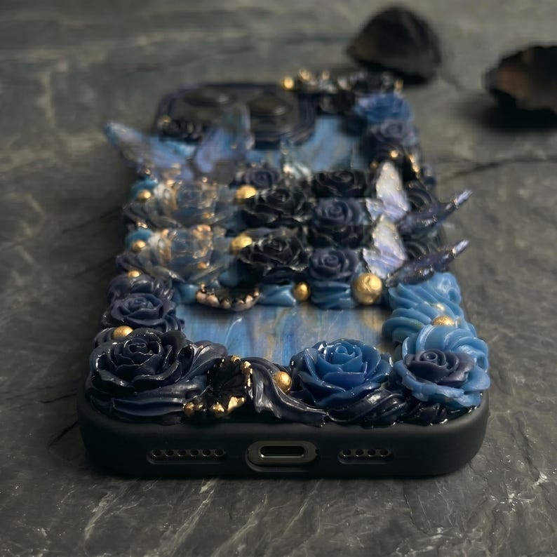 May include: A black phone case adorned with sculpted blue roses, butterflies, and gold accents. The case features a marbled blue background and is intricately detailed with a floral and butterfly design. The case has a glossy finish.