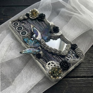 Puede incluir: Funda de teléfono decorativa con un diseño de vetas de madera oscura, adornada con mariposas, rosas negras y engranajes plateados. La funda presenta un pequeño espejo y un marco plateado. Las mariposas tienen alas iridiscentes azules y moradas.