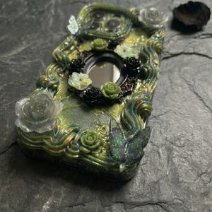 Op de afbeelding: Een decoratief telefoonhoesje met een groen en zwart harsoppervlak met wervelende texturen. De behuizing is versierd met driedimensionale elementen, waaronder kleine zwarte en groene rozen, een centrale ovale spiegel en twee vlindervormige accenten.