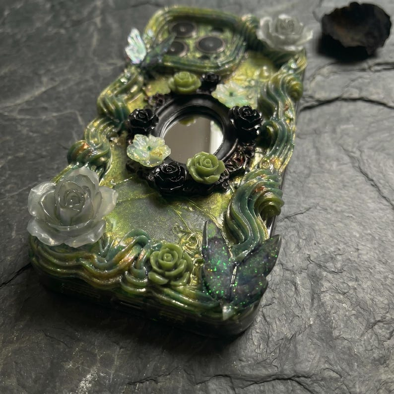 Op de afbeelding: Een decoratief telefoonhoesje met een groen en zwart harsoppervlak met wervelende texturen. De behuizing is versierd met driedimensionale elementen, waaronder kleine zwarte en groene rozen, een centrale ovale spiegel en twee vlindervormige accenten.