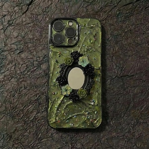 Op de afbeelding: Een smartphonehoesje met een textuurrijke olijfgroene basis, een centraal ovaal spiegelframe omringd door kleine zwarte en groene bloemversieringen en verspreide iriserende kralen, met een uitsparing voor de triple camera.