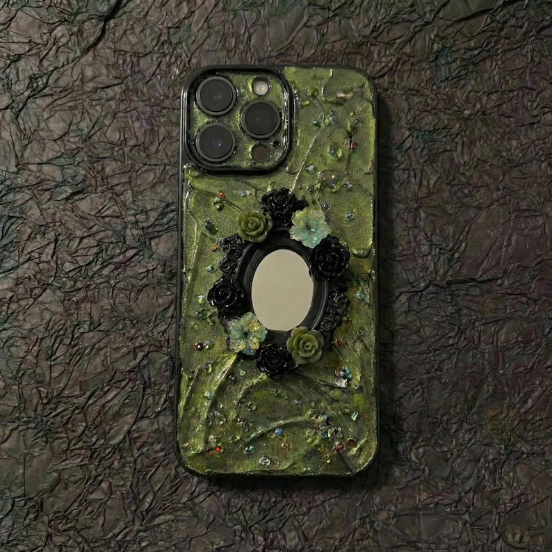 Op de afbeelding: Een smartphonehoesje met een textuurrijke olijfgroene basis, een centraal ovaal spiegelframe omringd door kleine zwarte en groene bloemversieringen en verspreide iriserende kralen, met een uitsparing voor de triple camera.