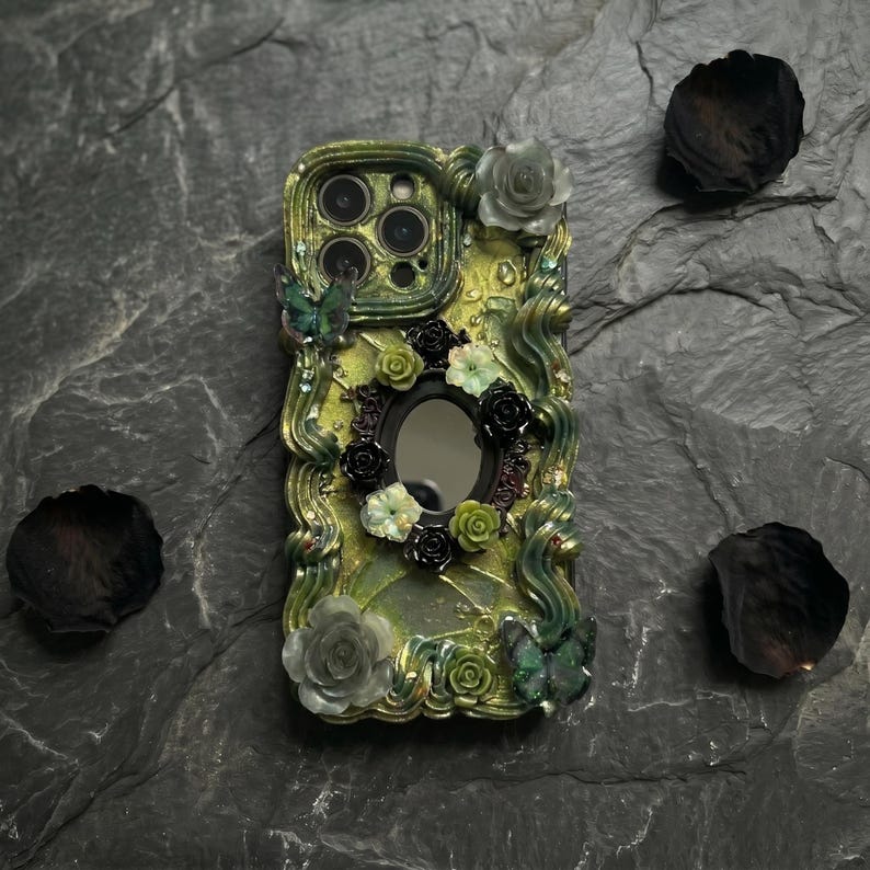 Op de afbeelding: Een telefoonhoesje met een textuur van olijfgroene en gouden basis met wervelende reli&euml;fpatronen. Het is versierd met kleine zwarte en groene bloemaccenten, twee vlinderfiguren en een centrale ovale spiegel in het ontwerp.